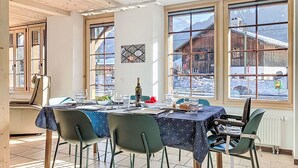 Uma sala de jantar com uma mesa posta para quatro pessoas, grandes janelas com vista para uma montanha nevada e um prédio de madeira, e um teto com vigas de madeira e um luminário moderno.