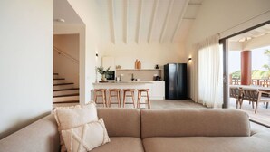 Ein modernes Wohnzimmer mit einem beigefarbenen Sofa, Holzschemeln, einer Küche mit einem schwarzen Kühlschrank und einem Essbereich mit Tisch und Stühlen.