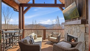 Eine überdachte Terrasse im Freien mit Korbmöbeln, einem Kamin aus Stein und Blick auf die Berge.