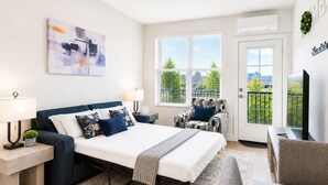 A modern bedroom with a bed, two chairs, a desk, and a window with a view of trees. - Modern 2BR Suite • Convention • Bathhouse Row (Hot Springs)