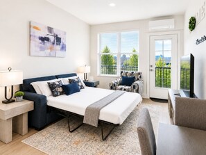 A modern bedroom with a bed, two chairs, a desk, and a window with a view of trees. - Modern 2BR Suite • Convention • Bathhouse Row (Hot Springs)