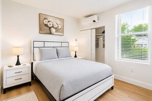 A bedroom with a bed, bedside tables, a nightstand, a wall-mounted air conditioner, a window with blinds, and a painting of flowers. - Modern 2BR Suite • Convention • Bathhouse Row (Hot Springs)