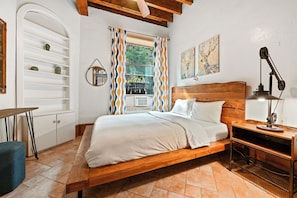 Un dormitorio con cama de madera, ropa de cama blanca, una mesita de noche con lámpara, una ventana con cortinas estampadas, un estante con objetos decorativos y un cuadro en la pared.