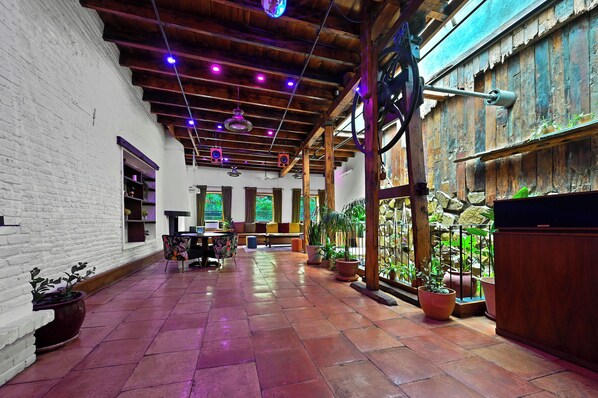 A spacious indoor area with a high ceiling, wooden beams, and terracotta flooring. There are multiple seating areas with cushions and tables, surrounded by potted plants.