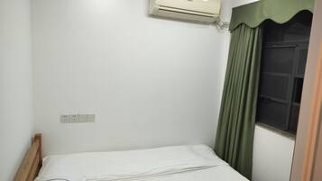 A small, clean room with a single bed, a window with green curtains, an air conditioning unit, and a power outlet on the wall.