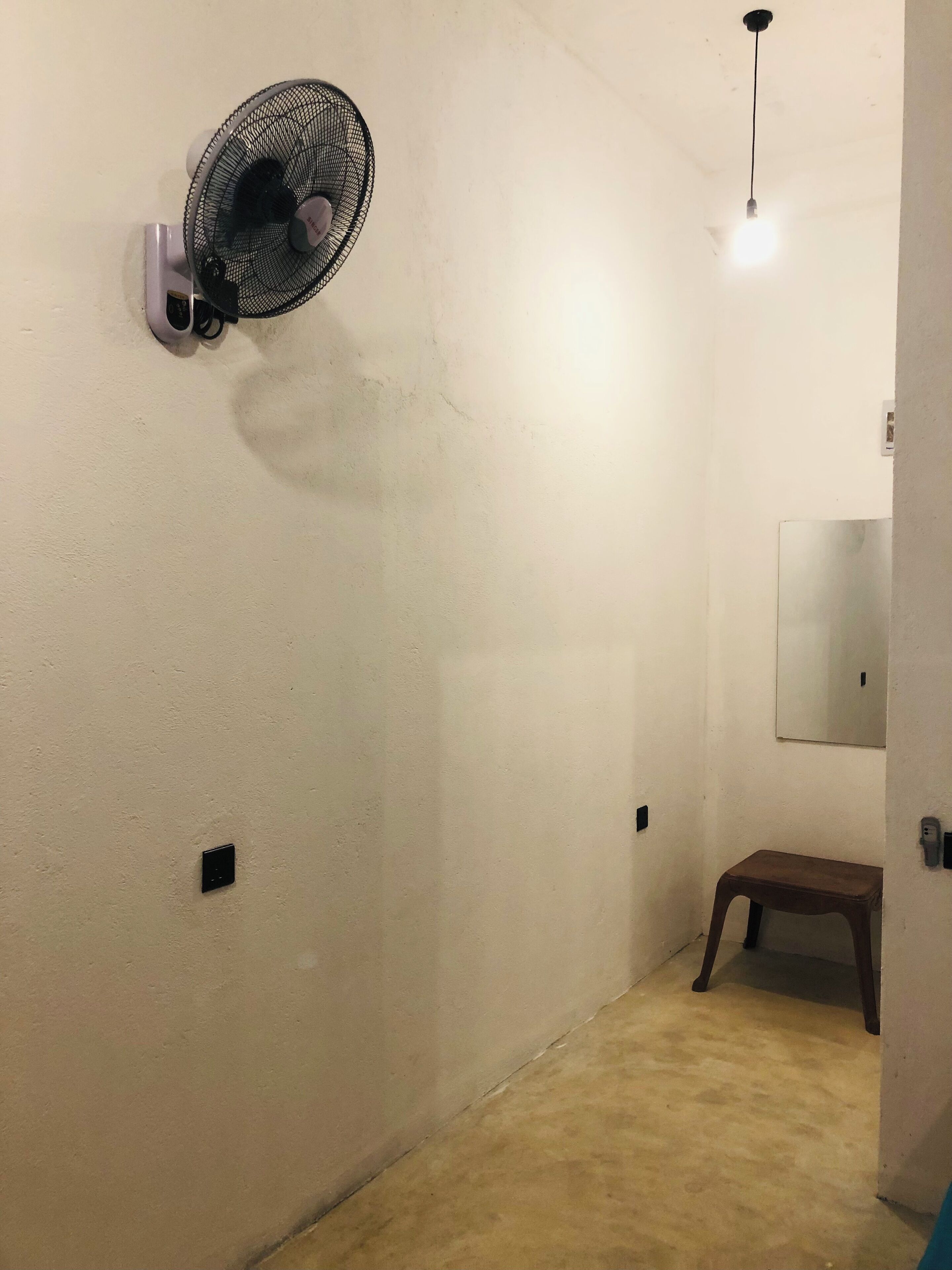 A hallway with a standing fan, a light fixture, a small stool, and a mirror on the wall.