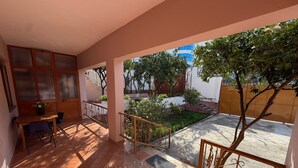 A sunlit patio with a wooden table and chairs, a small garden area, and a view of a building with blue windows.