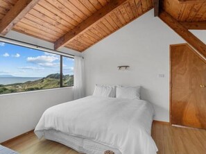 A bedroom with a large bed, a wooden ceiling, a window with a view of the sea, and a door. - Pakiri Beachhouse  Retreat and Relax (Tomarata)