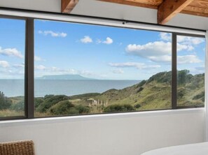 A room with a large window offering a view of the sea and hills. - Pakiri Beachhouse  Retreat and Relax (Tomarata)