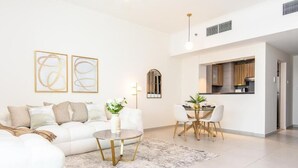 A modern living room with a white sofa, a round coffee table, and a dining area with a white table and chairs. There is a kitchen visible in the background. - Bloomfields 1Br Azure Stay Ajwan (Abu Dhabi)