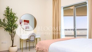 A bedroom with a bed, a desk, a chair, a potted plant, and a large window. - Bloomfields 1Br Azure Stay Ajwan (Abu Dhabi)