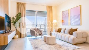 A modern living room with a sofa, a coffee table, and a TV on a wooden stand. There is a large window with curtains, a plant, and a painting on the wall. - Bloomfields 1Br Azure Stay Ajwan (Abu Dhabi)