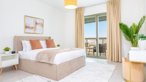 A modern bedroom with a large bed, a bedside table with a lamp, a window with curtains, and a view of the outdoors. - Bloomfields 1Br Azure Stay Ajwan (Abu Dhabi)