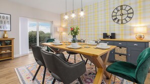 Un coin repas avec une table en bois dressée pour quatre personnes, une horloge au mur et une vue à travers une porte vitrée.
