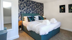 A neatly made bed with white linens and folded towels, a teal headboard, and a wall with a leaf pattern.