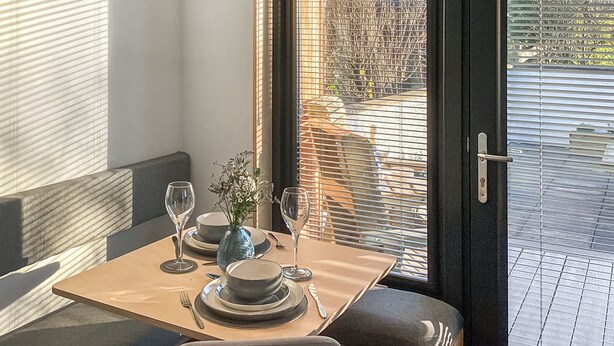 Un comedor con una mesa dispuesta para dos, un reloj en la pared y vistas a través de una puerta con persianas.