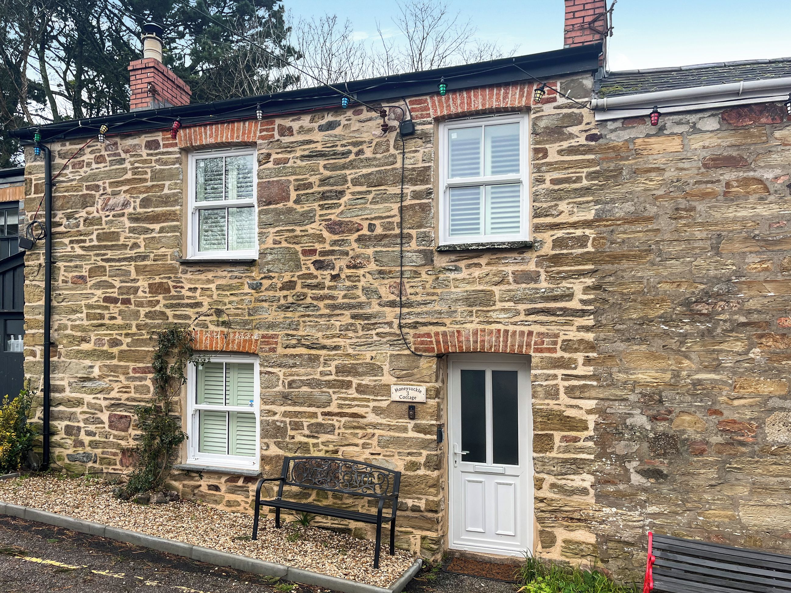 A stone cottage with a white door, two windows with shutters, a bench, and a sign reading "Honeysuckle Cottage".