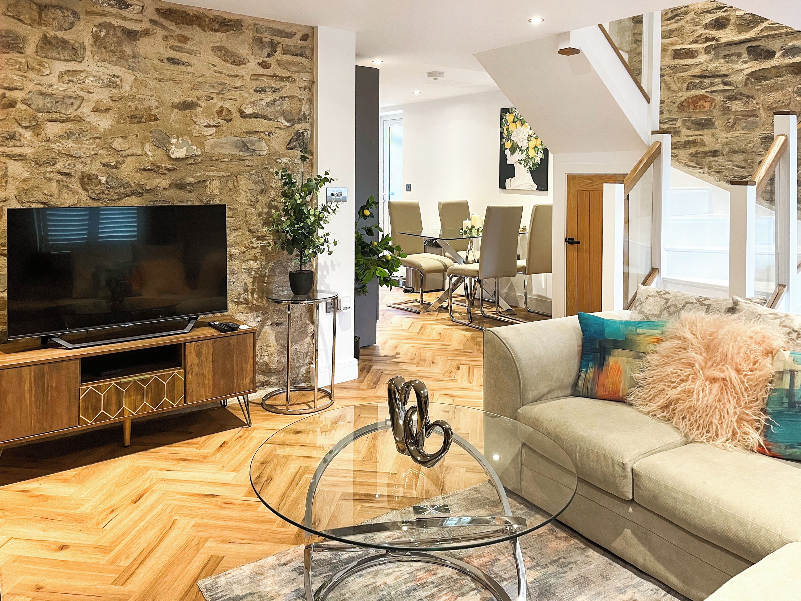 Un salon moderne avec un mur en pierre, un téléviseur à écran plat, une table basse en verre, un canapé beige et un coin repas avec des chaises et une table.