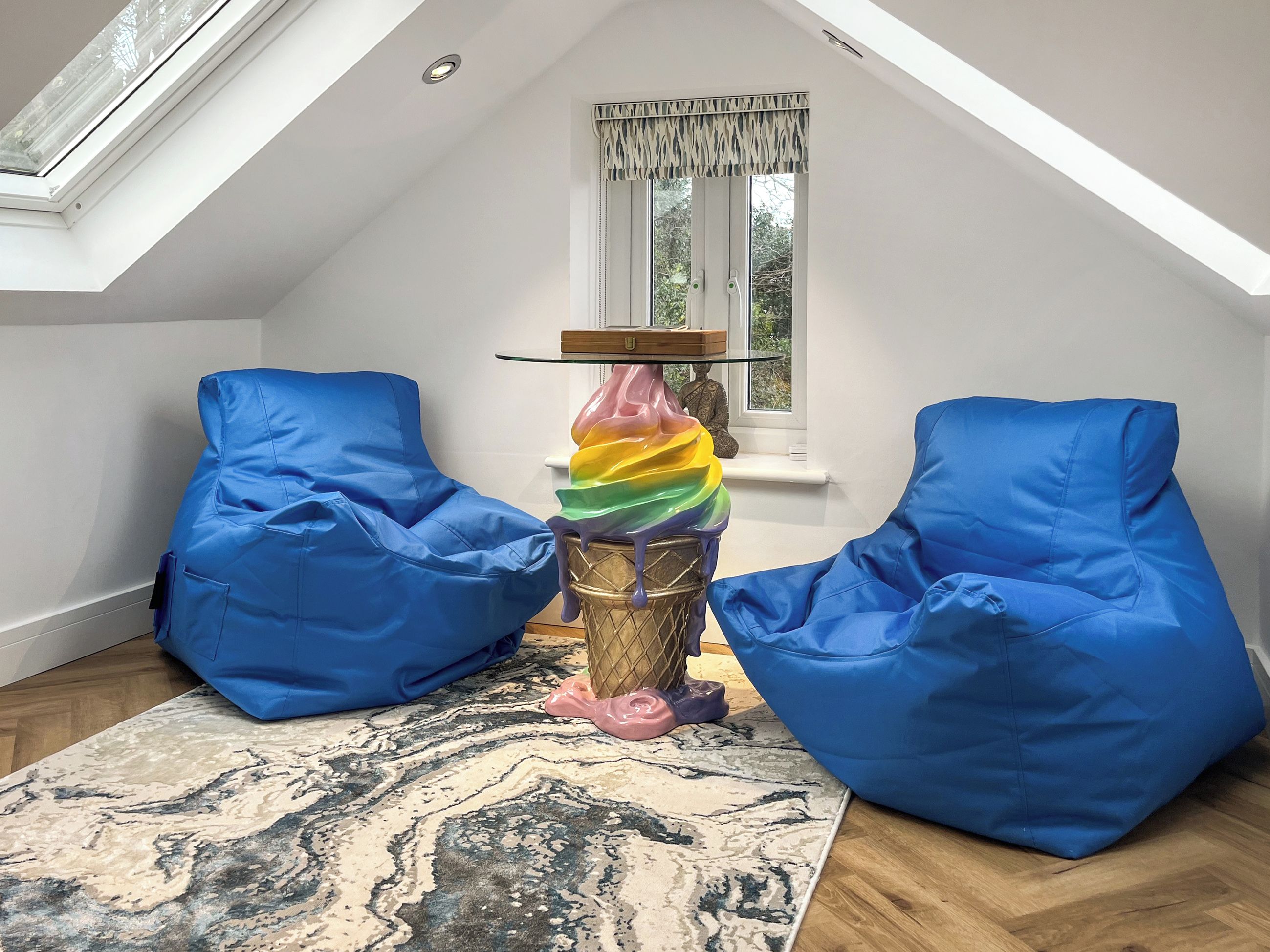A room with a sloped ceiling, a skylight, and a large window with a curtain. There are two blue bean bags and a colorful sculpture on a wooden table.