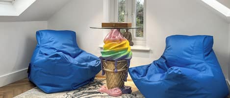 A room with a sloped ceiling, a skylight, and a large window with a curtain. There are two blue bean bags and a colorful sculpture on a wooden table.