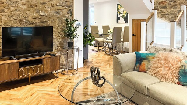 Ein modernes Wohnzimmer mit Steinwand, Flachbildfernseher, Glastisch, beiges Sofa und Essbereich mit Stühlen und Tisch.