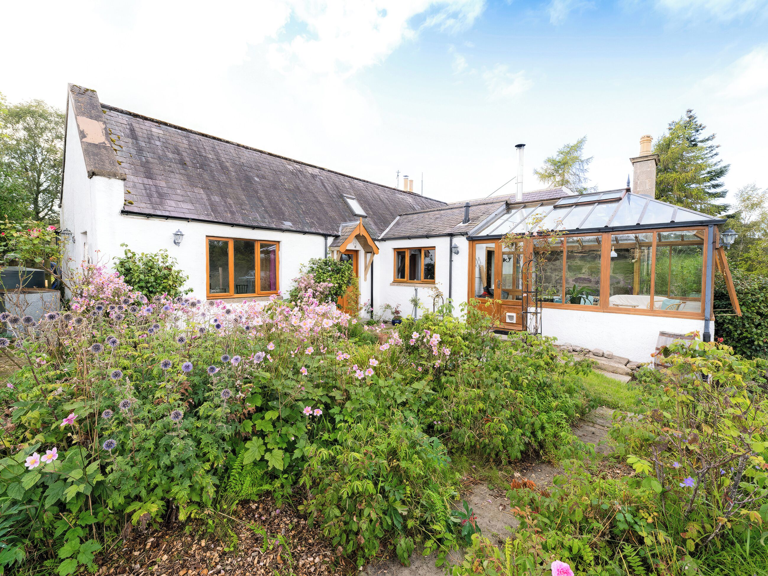 A white house with a large glass conservatory, surrounded by a garden with various plants and flowers.