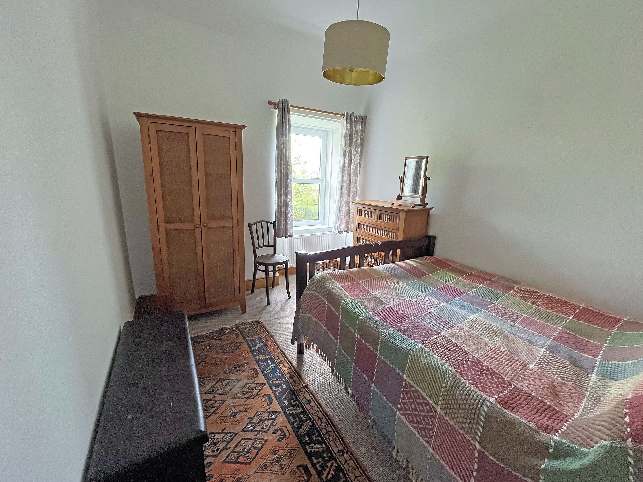 A bedroom with a bed, a wooden wardrobe, a desk with a chair, a hanging lamp, and a window with curtains.