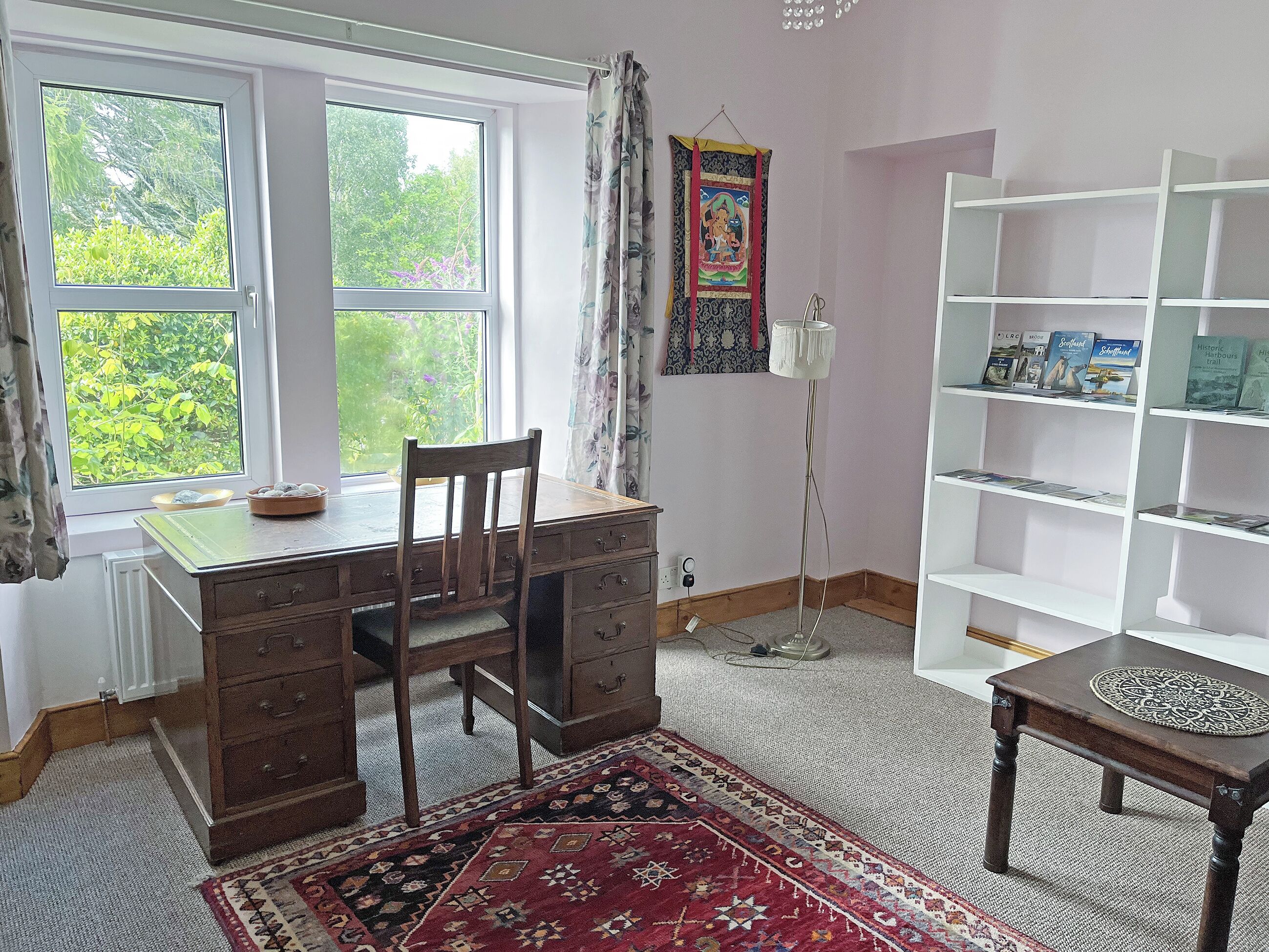 A room with a wooden desk, a chair, a bookshelf, a patterned rug, and a window with a view of greenery.