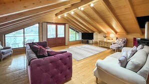 Habitación en el ático con techo de madera, un ventanal grande, un televisor y dos sofás.
