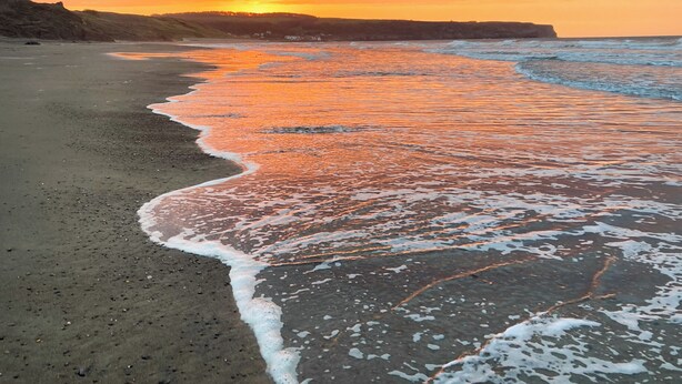 Una playa con olas, un atardecer y colinas al lejano.