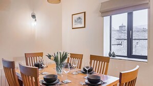 En matplats med ett träbord dukat för fyra personer, stolar, en vas med blommor, en inramad tavla på väggen och ett fönster med utsikt över byggnader.