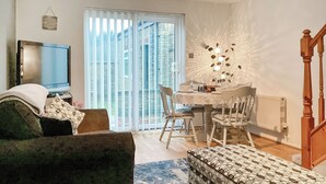 Una sala de estar con sofá, un reposapiés estampado, una mesa de comedor con sillas, televisor y puerta corrediza de vidrio que da a un balcón.