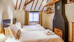 Una cama doble con sábanas blancas, una mesita de noche con lámpara, una ventana con cortinas, una chimenea con repisa y un techo de madera.