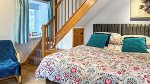 Une chambre avec un escalier en bois, un lit avec une literie à motifs floraux, un fauteuil bleu et une fenêtre avec des rideaux.