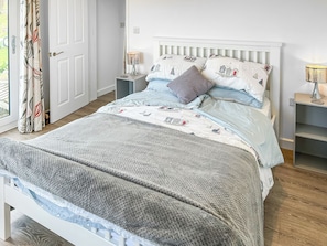 A neatly made bed with a patterned bedspread, a grey knitted throw, and matching pillows. There is a bedside table with a lamp, a window with curtains, and a door. - Escape to something special, surrounded by peaceful countryside. (St Just)