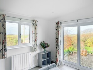 A room with a radiator, a grey sofa, a small table with a vase, and a view of a garden through a window. - Escape to something special, surrounded by peaceful countryside. (St Just)