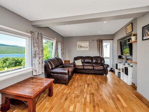 Ein modernes Wohnzimmer mit einem Ledersofa, Holzboden, einem Fernseher und Blick auf einen See.