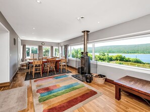 A modern interior with a dining area, a large window with a view of a lake, and a wood-burning stove. - Overscaig House - UK51360 (Lairg)