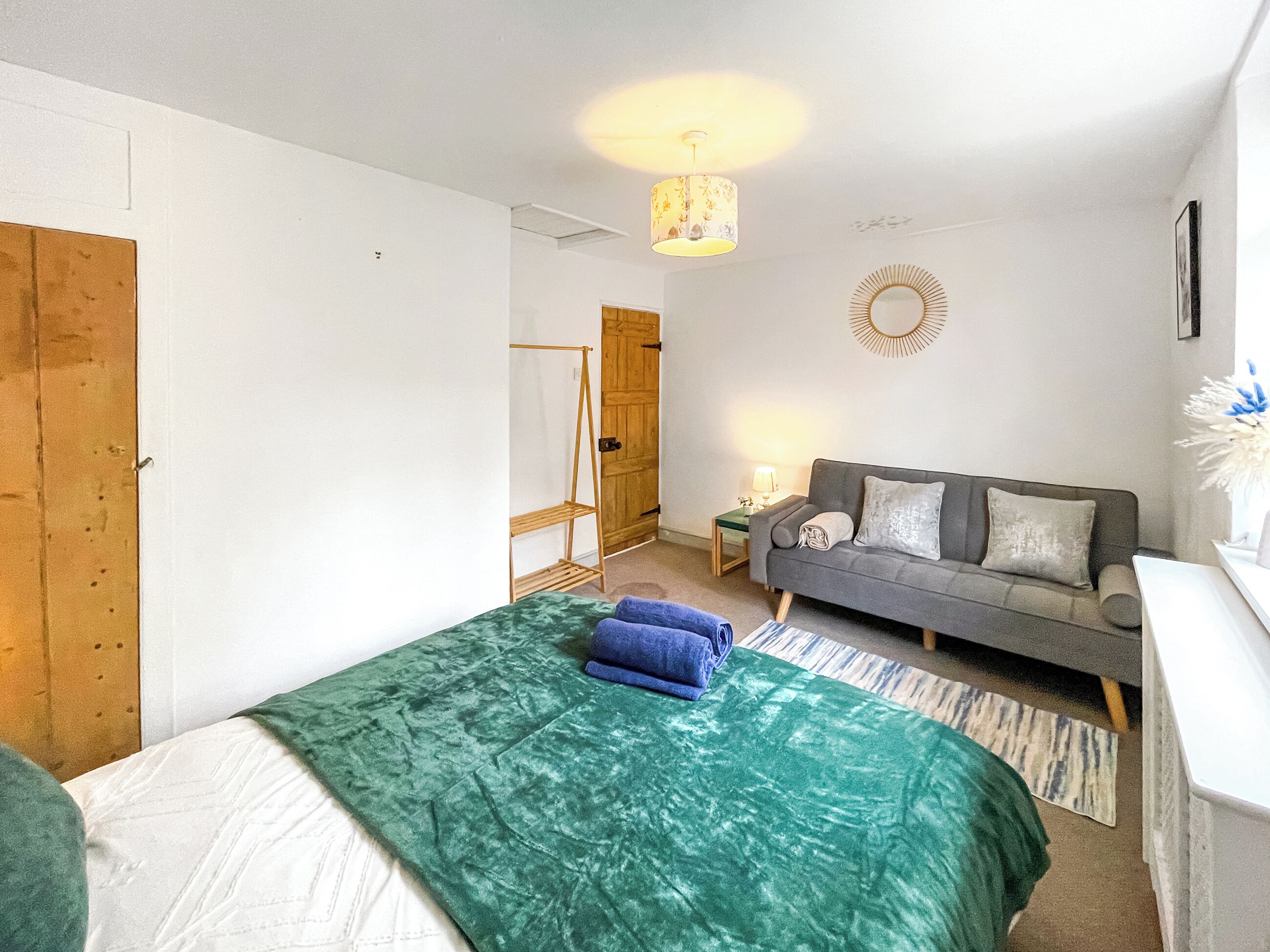 A room with a bed, a grey sofa, a small table with a lamp, and a wooden wardrobe.
