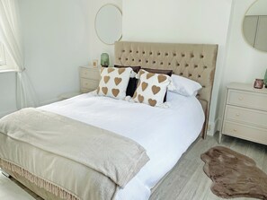 A neatly made bed with heart-patterned pillows, a tufted headboard, a bedside table with a green lamp, and a round mirror on the wall. - 2 bedroom accommodation in Broadstairs (Broadstairs)