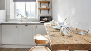 Una cocina con una mesa de madera, armarios blancos, un ventanal con persiana azul y un microondas en la encimera.