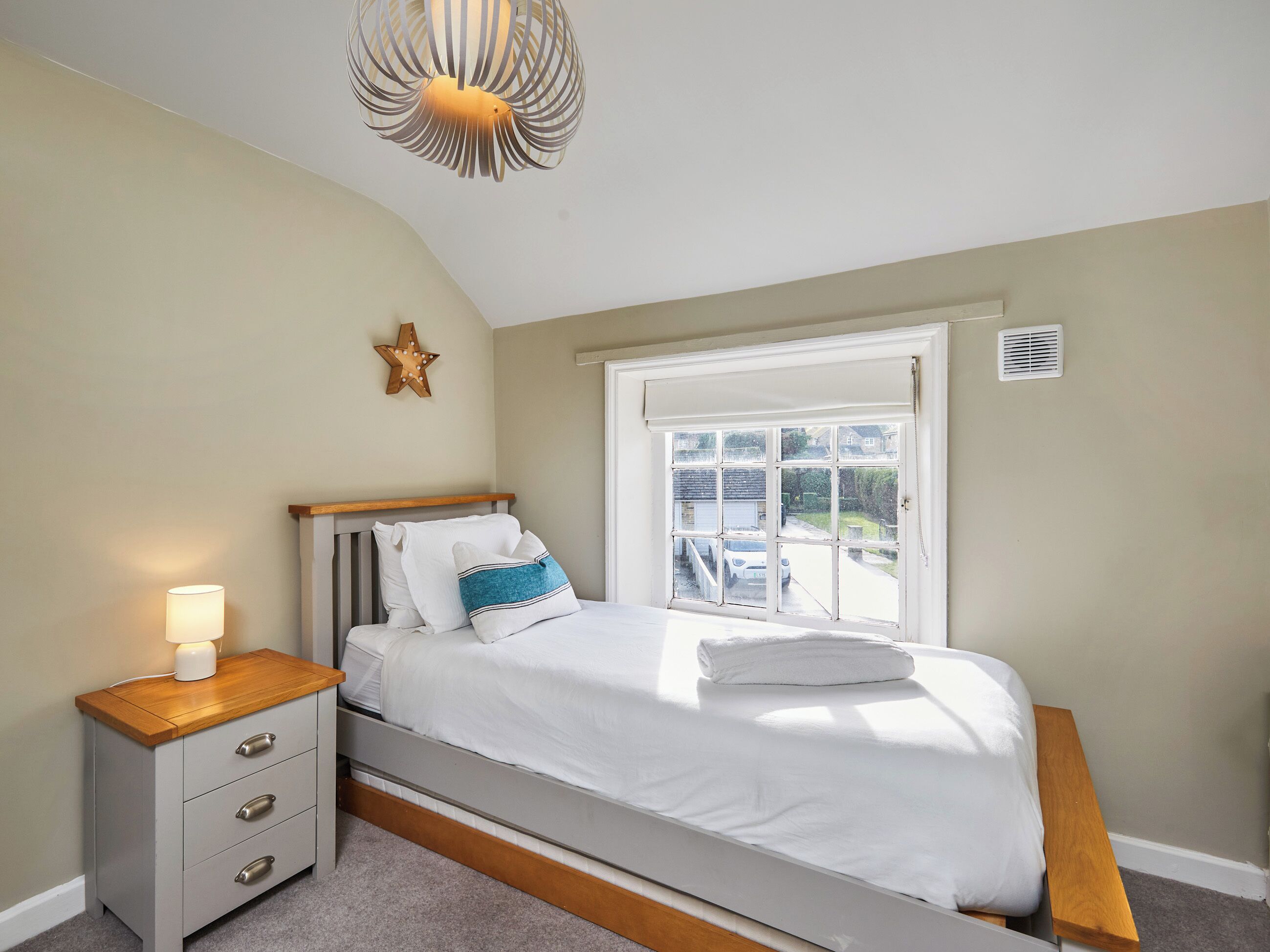 A bedroom with a bed, bedside table, lamp, star decoration, window, and a view of the outside.