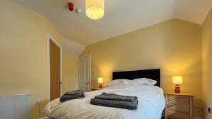 Una cama bien hecha con sábanas blancas y toallas dobladas, dos mesitas de noche con lámparas, una puerta y un radiador en la pared.