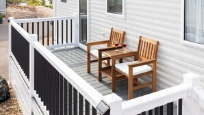 Uma casa móvel branca com um deck de madeira, duas cadeiras de madeira e uma mesa com dois copos de vinho tinto.
