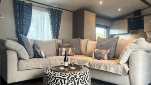A modern living room with a beige sectional sofa, patterned cushions, a round ottoman, and a small table with two wine glasses and a bottle. - Herdwick Lodge - UK51383 (Brocklebank, near Caldbeck and Uldale)