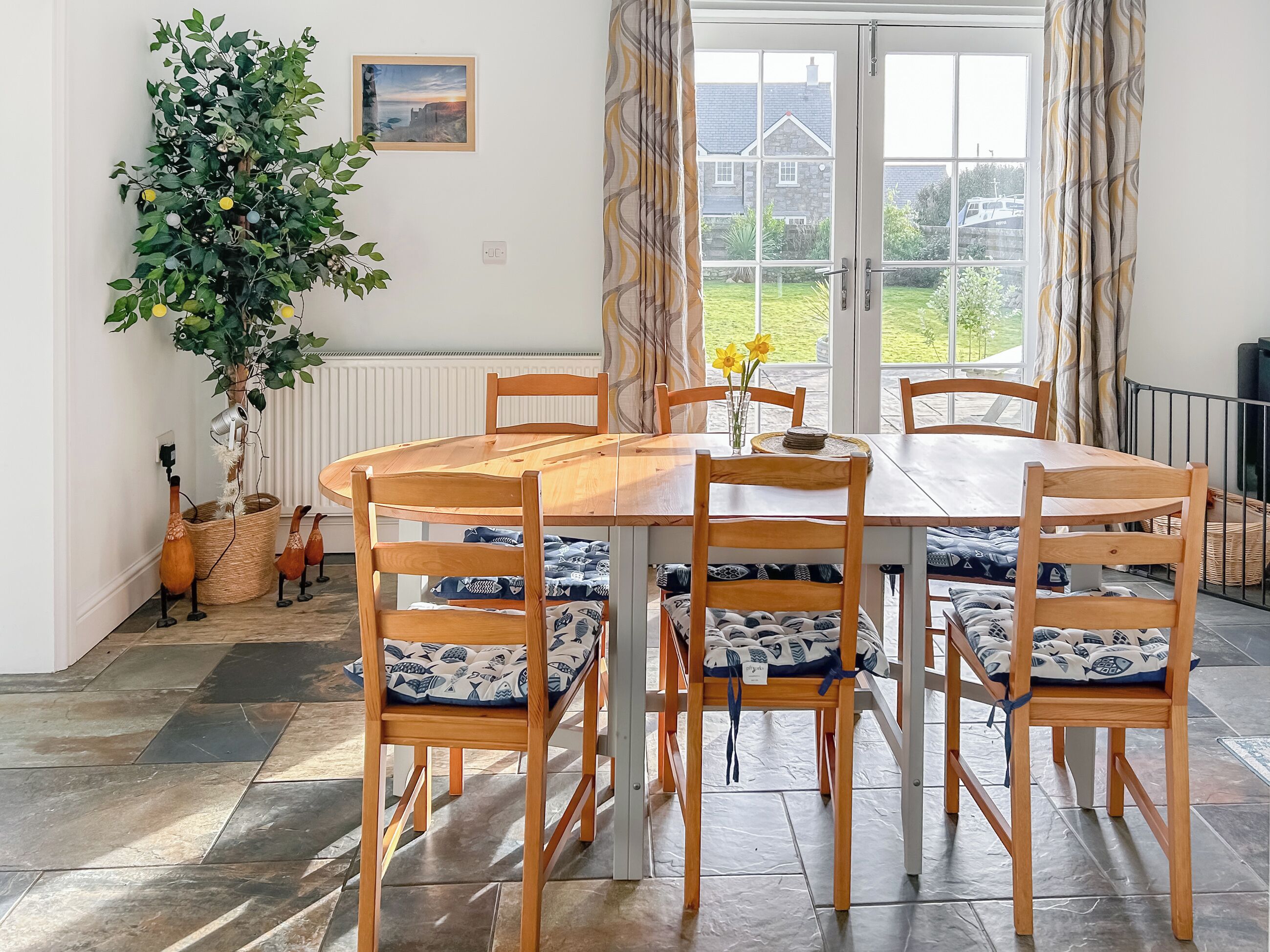 A dining area with a round wooden table, six wooden chairs with patterned cushions, a potted plant, and a view of the garden through a glass door.