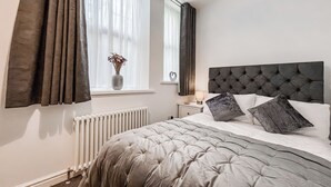 Une chambre à coucher avec un lit, une table de chevet, une fenêtre avec des rideaux, un radiateur et un vase avec des fleurs.