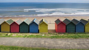 Une rangée de cabanes de plage aux couleurs vives longe une route côtière, avec l’océan en arrière-plan.