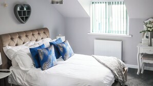 Ein Schlafzimmer mit einem karottenbesetzten Kopfteil, weißer Bettwäsche, blau gemusterten Kissen, einer Hängeleuchte, einem Heizkörper, einem Fenster mit Vorhängen und einem Nachttisch mit einer Vase.