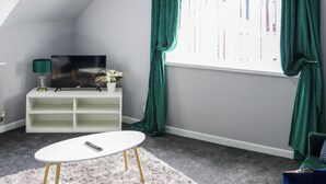 Ein Zimmer mit Fernseher auf einem weißen Schrank, einem kleinen weißen Tisch, einem flauschigen Teppich und grünen Vorhängen.